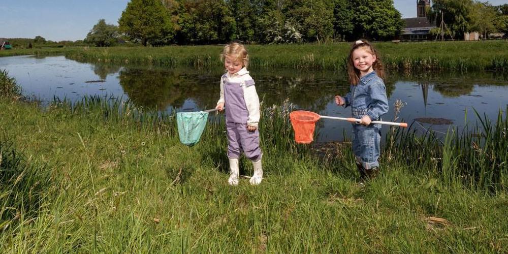 Twee jonge kinderen vangen waterdiertjes met schepnetten langs de oever van een sloot, met een kerktoren en groene bomen op de achtergrond.