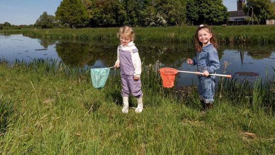 Twee jonge kinderen vangen waterdiertjes met schepnetten langs de oever van een sloot, met een kerktoren en groene bomen op de achtergrond.