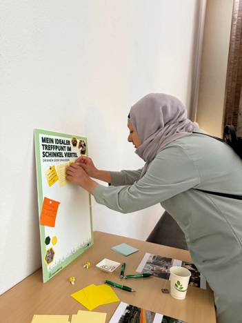 Een vrouw plakt een gele post-it op een bord met de tekst "Mein idealer Treffpunkt im Schinkel Viertel" tijdens een participatie-evenement.