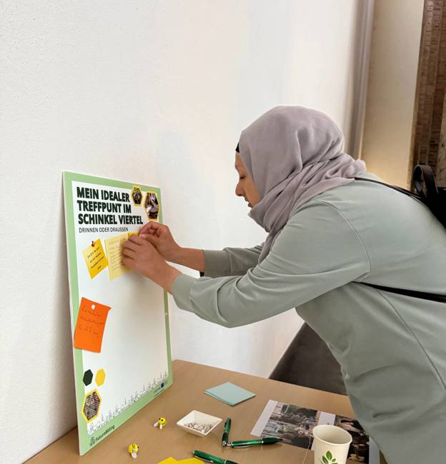 Een vrouw plakt een gele post-it op een bord met de tekst "Mein idealer Treffpunkt im Schinkel Viertel" tijdens een participatie-evenement.