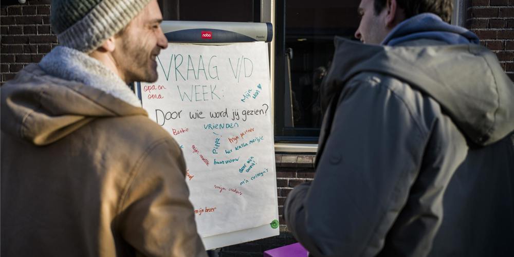 Twee mannen staan voor een flip-over waarop staat geschreven 'Vraag v/d week. Door wie word jij gezien?'