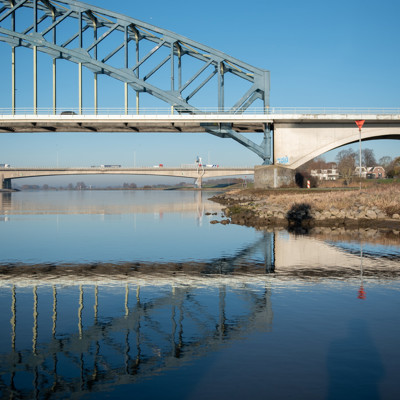 Twee bruggen over een brede rivier op een heldere dag, met hun reflectie zichtbaar in het rustige water.