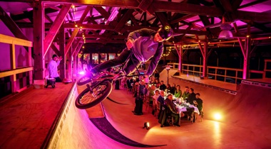 Foto in skatehall waarbij een stuntfietser in actie te zien in van dichtbij. Op de achtergrond staat een lange dinertafel opgesteld waaraan gasten zitten. Zij kijken naar de stuntfietser.
