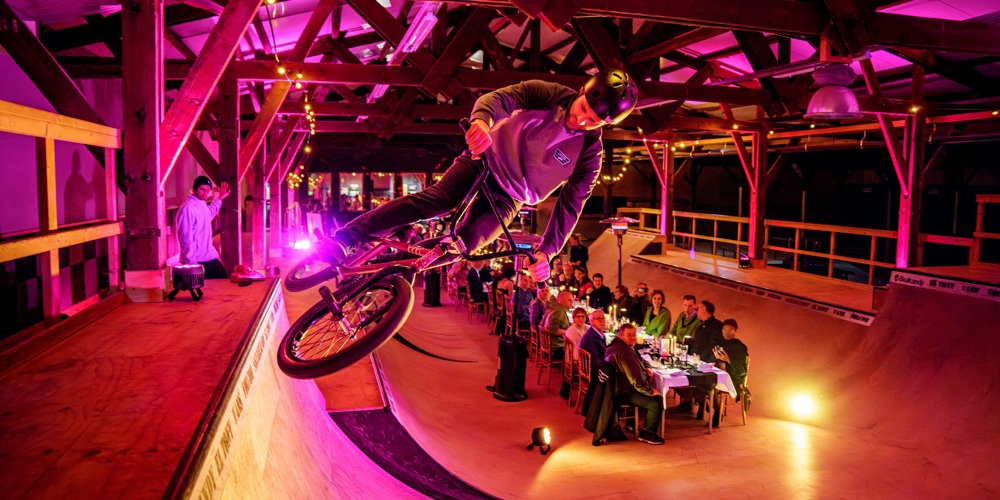 Foto in skatehall waarbij een stuntfietser in actie te zien in van dichtbij. Op de achtergrond staat een lange dinertafel opgesteld waaraan gasten zitten. Zij kijken naar de stuntfietser.