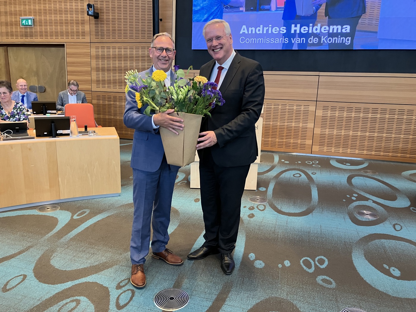 Spiker en de Commissaris van de Koning (CdK) in de Statenzaal. Spiker heeft een bos bloemen vast. Op de achtergrond zien we ook enkele gedeputeerden zitten. En een stukje van het grote presentatiescherm in de Statenzaal met daarop de naam en functie van de CdK.