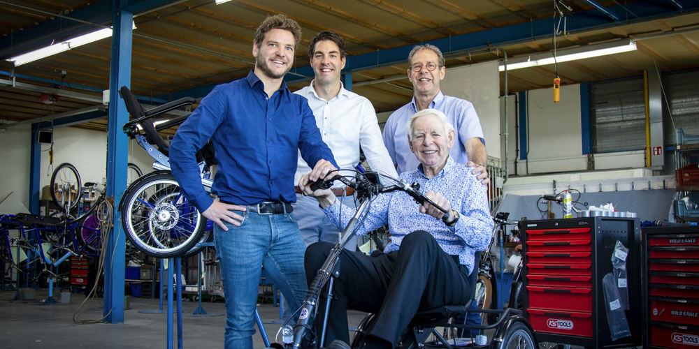 Drie mensen staan om een man die zit op een driewieler in een fietsenmakerswinkel