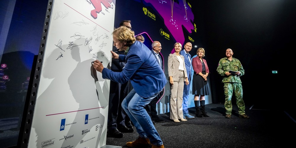 Een groep mensen poseert op een podium voor een groot scherm met de tekst “Intentieverklaring Regionale Innovatiesamenwerking” en een kaart van Nederland met logo’s van regionale ontwikkelingsmaatschappijen.