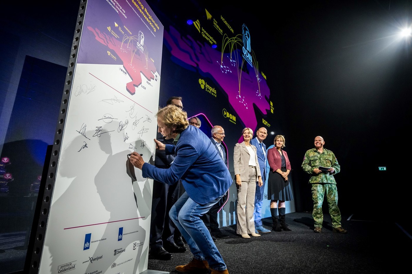 Een groep mensen poseert op een podium voor een groot scherm met de tekst “Intentieverklaring Regionale Innovatiesamenwerking” en een kaart van Nederland met logo’s van regionale ontwikkelingsmaatschappijen.