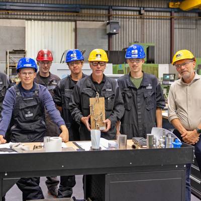 Groep werknemers met helm poseert in fabriek, één persoon houdt een trofee vast.