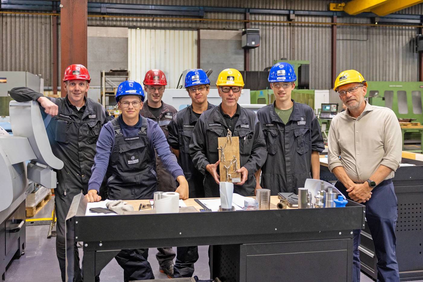 Groep werknemers met helm poseert in fabriek, één persoon houdt een trofee vast.
