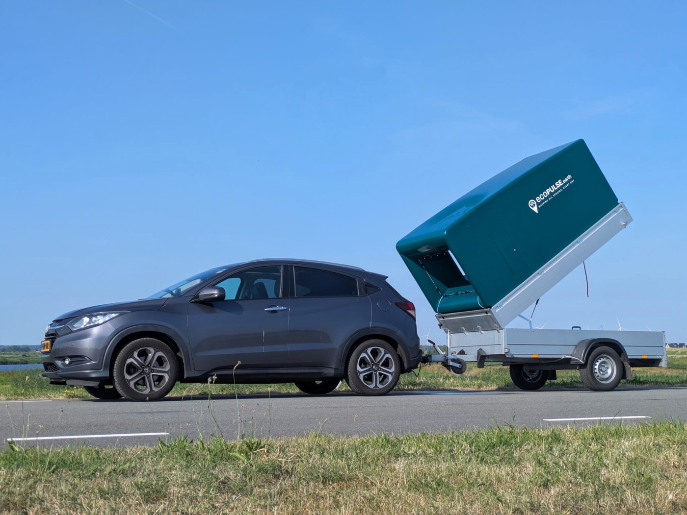 Een auto met een aanhanger van EcoPulse staat buiten stil langs de weg. De bovenkant van de aanhanger staat in een hoek van ongeveer 45 graden omhoog om lucht op te kunnen vangen en analyseren.