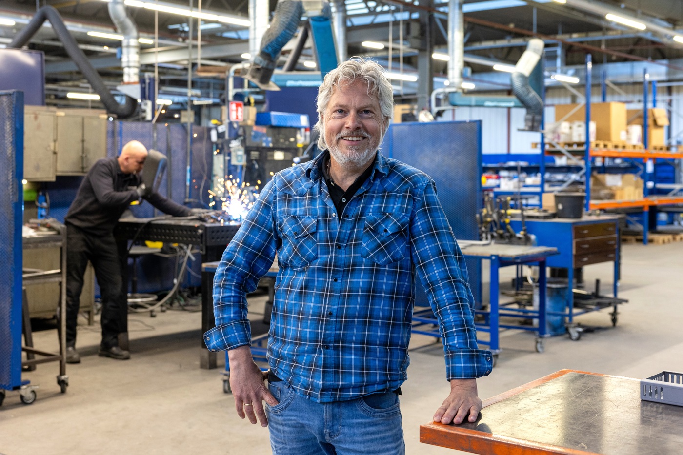 Portret in de werk en leeromgeving met Henri vooraan in een blauw-groen geruite blouse. Op de achtergrond in de werkplaats machines en iemand die aan het lassen is.