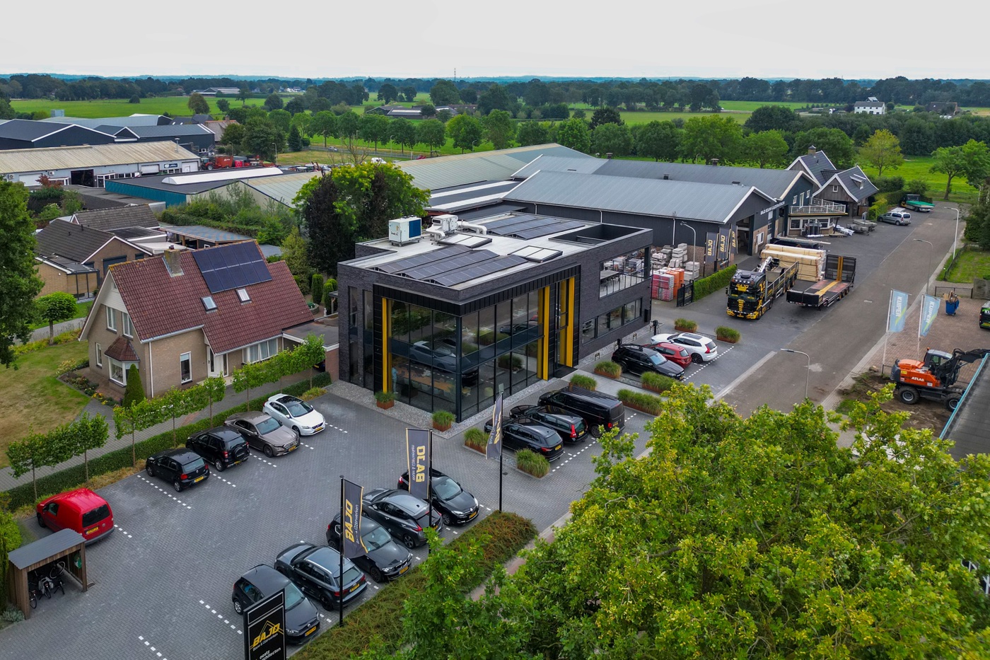 Het bedrijf vanuit de lucht gezien, op het industrieterrein. Het is overdag. Buiten staan auto's geparkeerd. Naast het kantoorgebouw wordt bij een bedrijfshal van het bouwbedrijf een vrachtwagen met daarnaast een grote lading hout voor een bouwproject.  