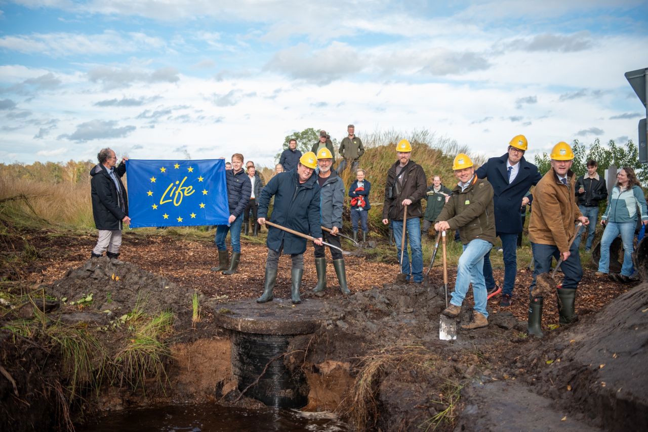 De start door zes mensen met een schop in de hand en een gele helm op. Rechts in beeld tonen 2 mensen de blauwe EU-vlag met de tekst 'Life'. Op de achtergrond kijken mensen naar de starthandeling.