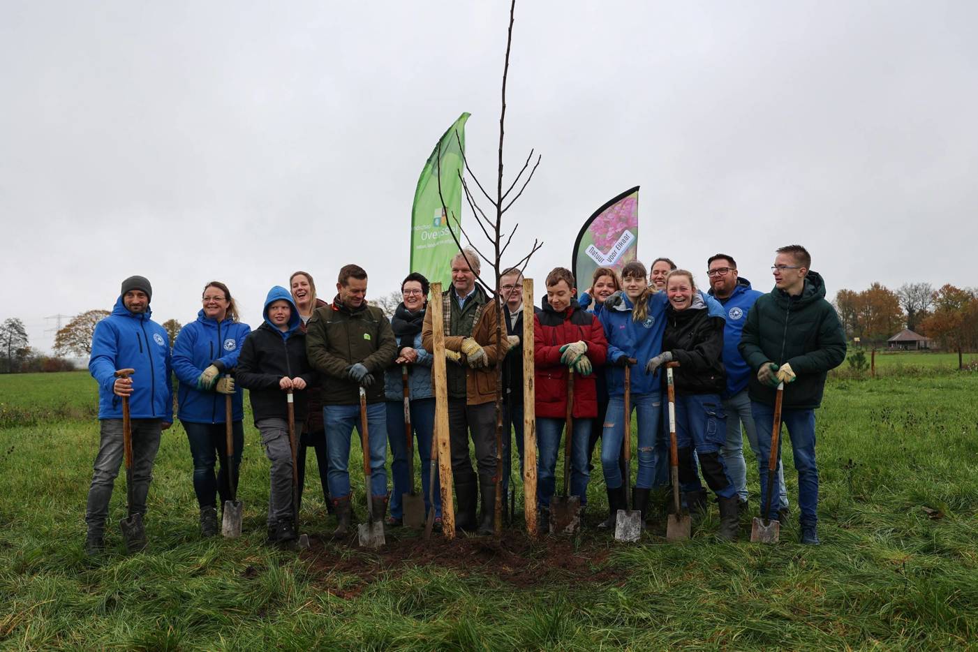 Groep mensen staat samen rondom een pas geplante boom met schoppen in de handen, glimlachend voor een foto.