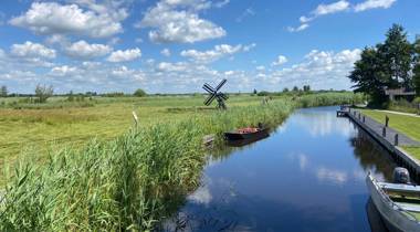 Rustig waterkanaal met rietkragen, kleine bootjes en een houten poldermolentje in natuurgebied Wieden Weerribben onder een blauwe lucht met wolken.