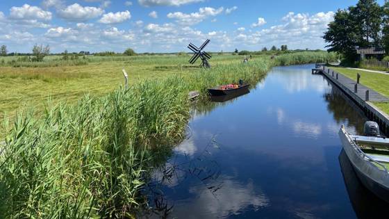 Rustig waterkanaal met rietkragen, kleine bootjes en een houten poldermolentje in natuurgebied Wieden Weerribben onder een blauwe lucht met wolken.