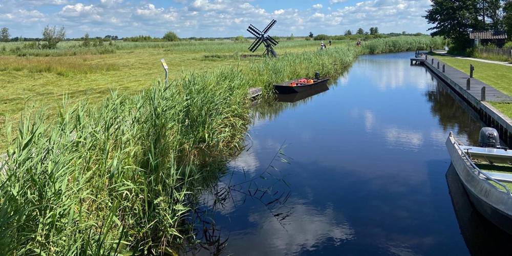 Rustig waterkanaal met rietkragen, kleine bootjes en een houten poldermolentje in natuurgebied Wieden Weerribben onder een blauwe lucht met wolken.