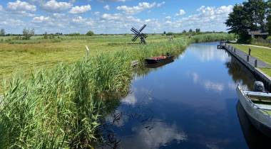 Rustig waterkanaal met rietkragen, kleine bootjes en een houten poldermolentje in natuurgebied Wieden Weerribben onder een blauwe lucht met wolken.